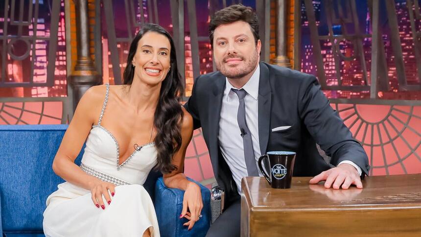 Sheilla Castro de vestido branco, sorrindo, com a mão no braço do sofá azul ao lado de Danilo Gentili durante gravação do The Noite, do SBT