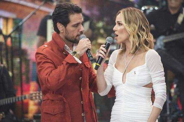 Thiago Aracam de jaqueta vermelha segurando o microfone ao lado de Ana Furtado, de vestido vermelho cantando com Thiago durante gravação do Faustão na Band