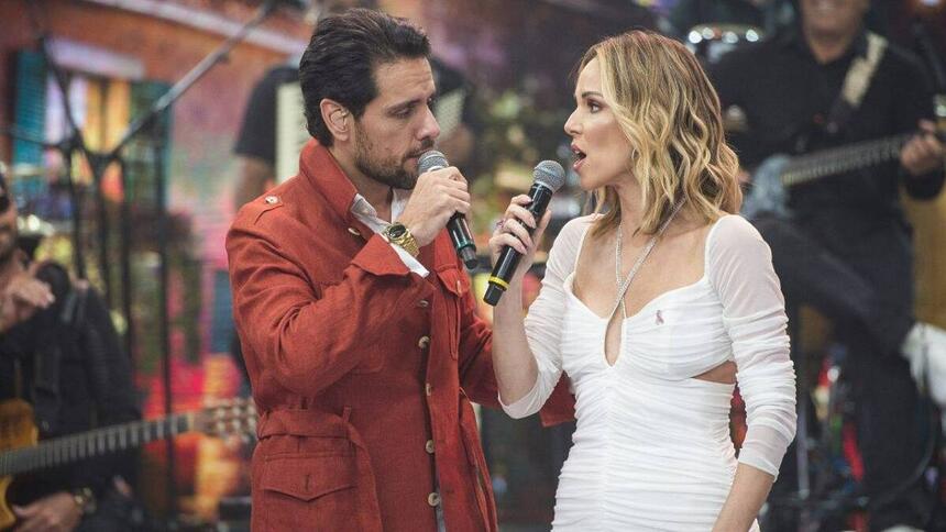 Thiago Aracam de jaqueta vermelha segurando o microfone ao lado de Ana Furtado, de vestido vermelho cantando com Thiago durante gravação do Faustão na Band