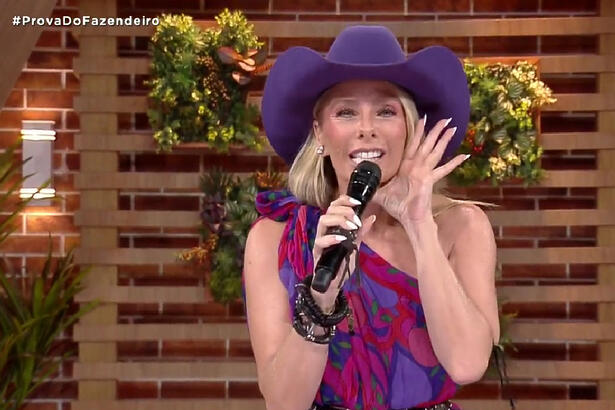 Adriane Galisteu de chapéu roxo, com a mão aberta na boca, segurando um microfone durante apresentação de A Fazenda, da Record TV