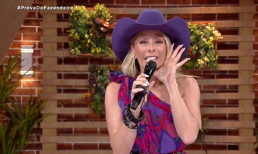 Adriane Galisteu de chapéu roxo, com a mão aberta na boca, segurando um microfone durante apresentação de A Fazenda, da Record TV