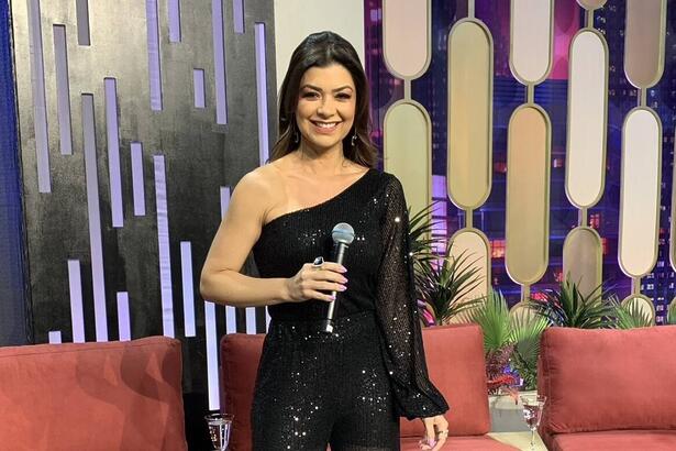 Amanda Françozo de vestido preto, sorrindo, segurando o microfone com o braço direito durante gravação do De Papo com Amanda Françozo, da TV Aparecida