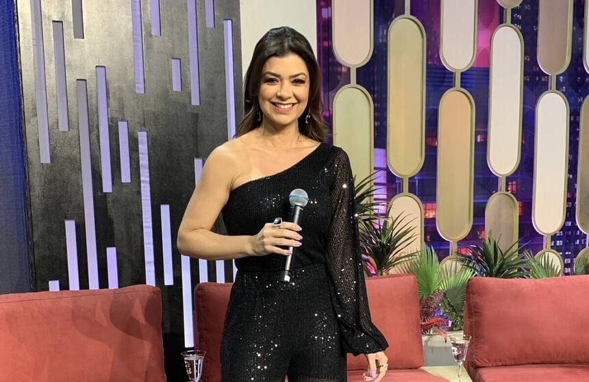 Amanda Françozo de vestido preto, sorrindo, segurando o microfone com o braço direito durante gravação do De Papo com Amanda Françozo, da TV Aparecida