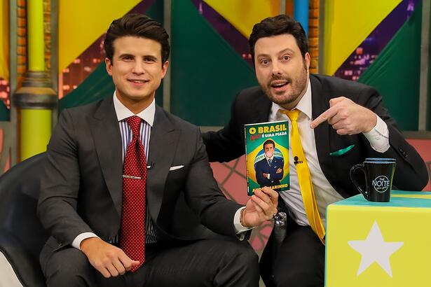 André Marinho de palitó preto, gravata vermelha, sentado, sorrindo, ao lado de Danilo Gentili durante gravação do The Noite, do SBT