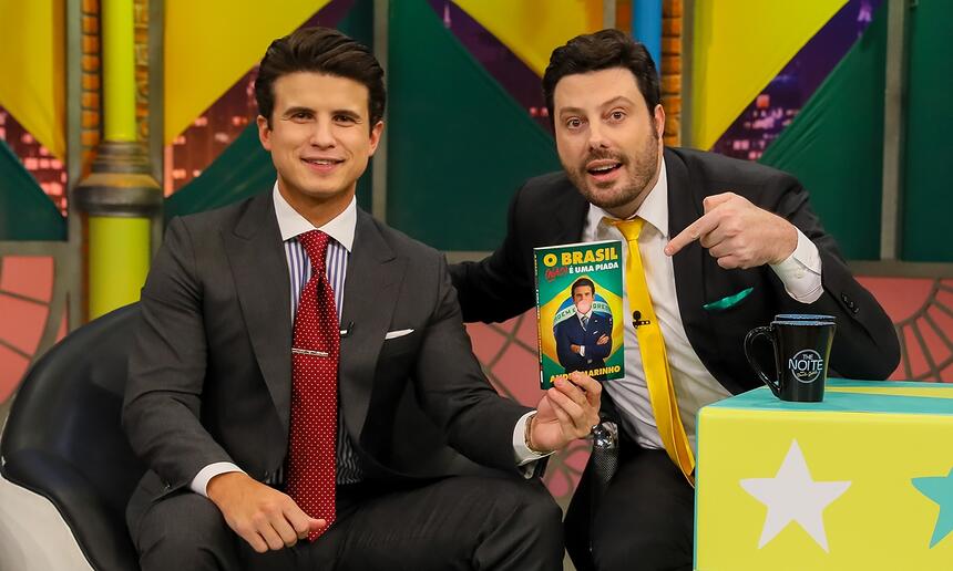 André Marinho de palitó preto, gravata vermelha, sentado, sorrindo, ao lado de Danilo Gentili durante gravação do The Noite, do SBT