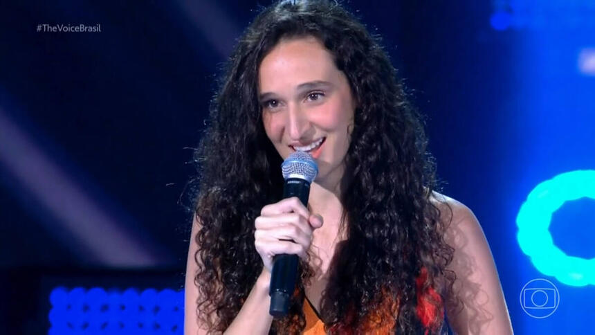 Antonia Medeiros cantando no palco do The Voice Brasil, com vestido florido e microfone na mão