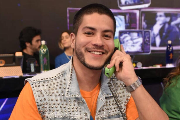 Arthur Aguiar com uma blusa laranja e um colete jeans por cima. Ele está com um sorriso aberto e com um telefone verde no ouvido, sentado no mesão da esperança