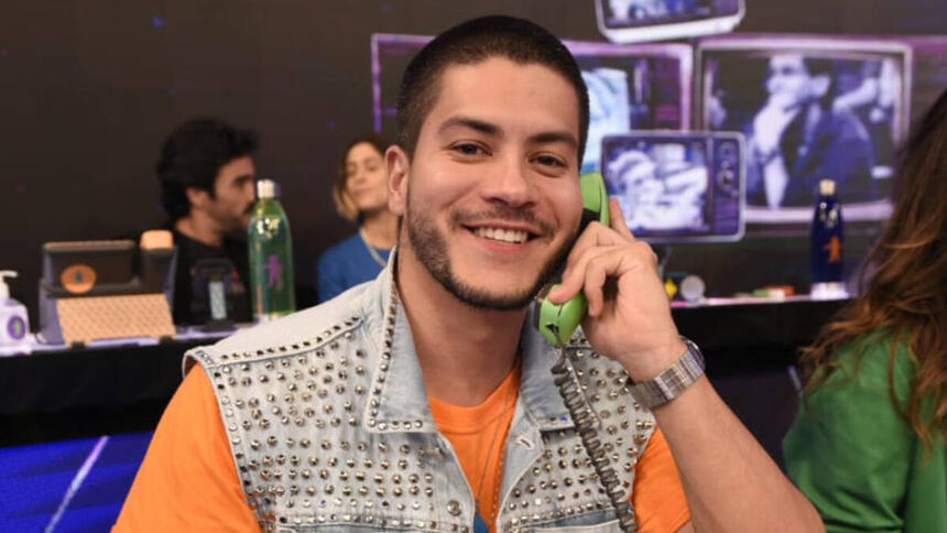 Arthur Aguiar com uma blusa laranja e um colete jeans por cima. Ele está com um sorriso aberto e com um telefone verde no ouvido, sentado no mesão da esperança