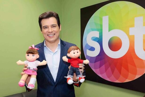 Celso Portiolli segurando os mascotes do Teleton, em uma sala, com o logo do SBT ao fundo