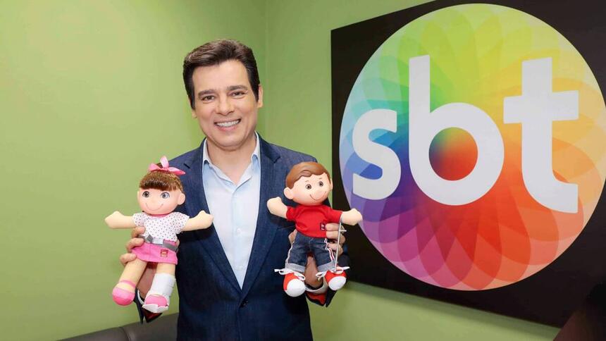 Celso Portiolli segurando os mascotes do Teleton, em uma sala, com o logo do SBT ao fundo