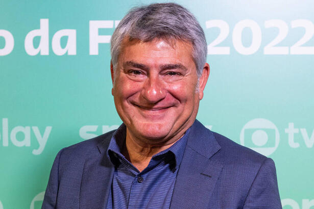 Cléber Machado com um sorriso no jogo, em um terno mais escuro, em frente a um banner azul da Globo