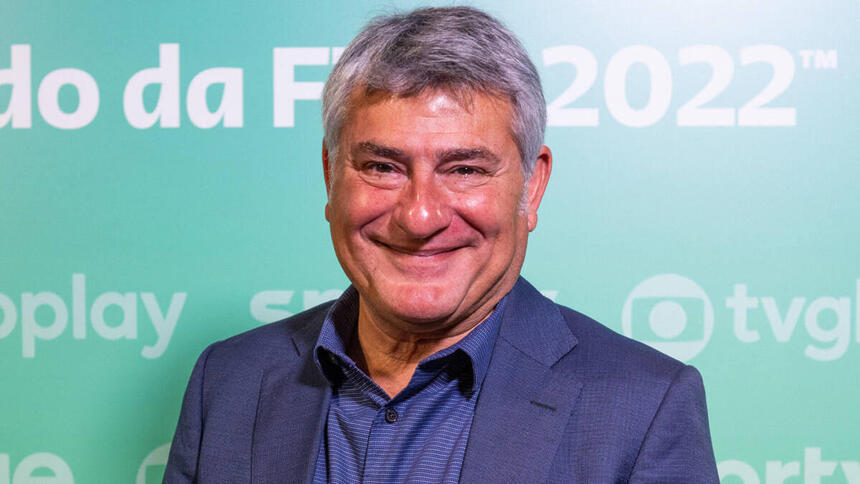 Cléber Machado com um sorriso no jogo, em um terno mais escuro, em frente a um banner azul da Globo