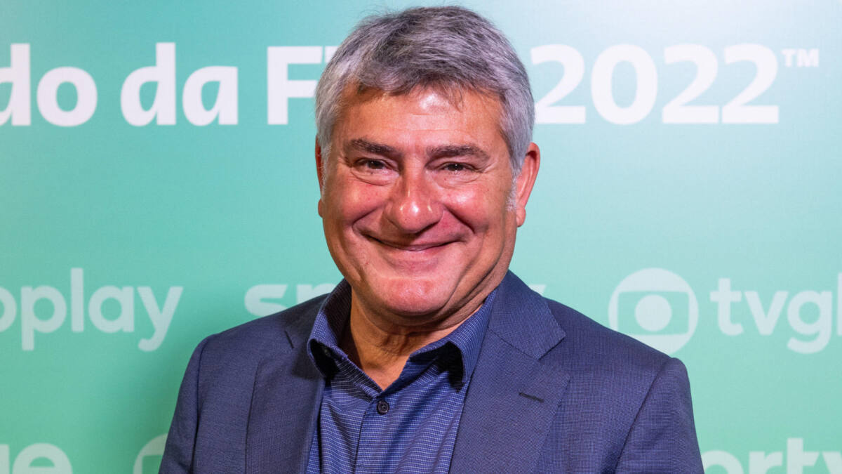 Cléber Machado com um sorriso no jogo, em um terno mais escuro, em frente a um banner azul da Globo