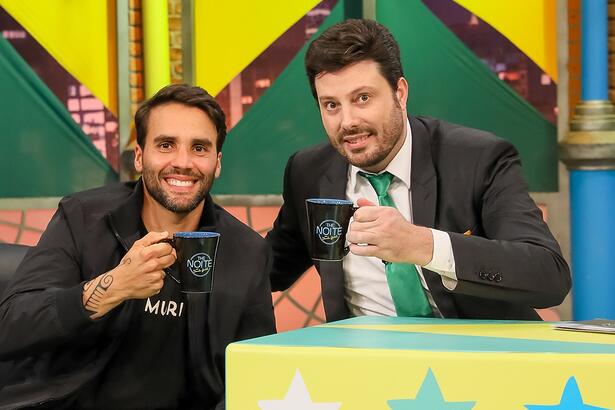 Daniel Candy sorrindo, de camisa preta, sentado num sofá durante gravação do The Noite, do SBT, ao lado de Danilo Gentili