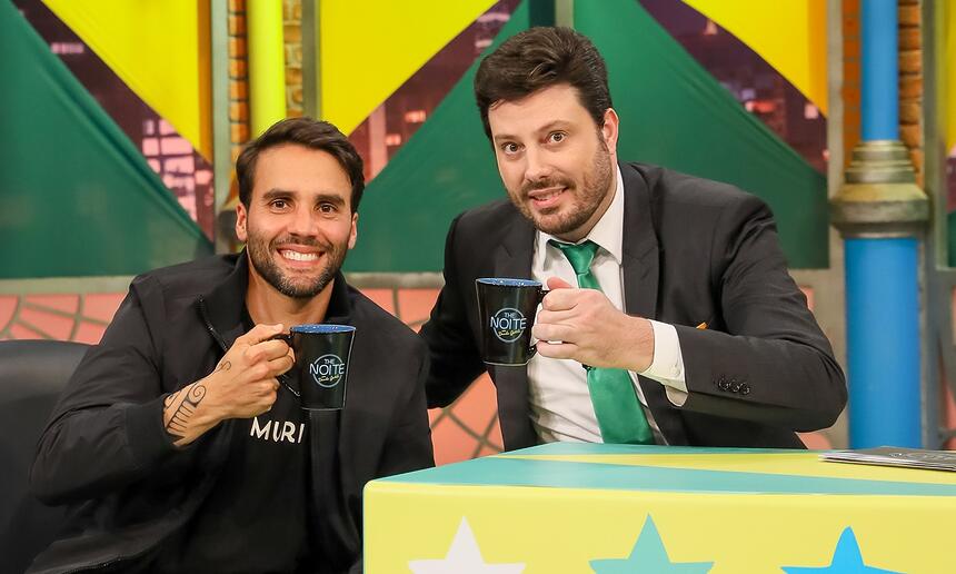 Daniel Candy sorrindo, de camisa preta, sentado num sofá durante gravação do The Noite, do SBT, ao lado de Danilo Gentili