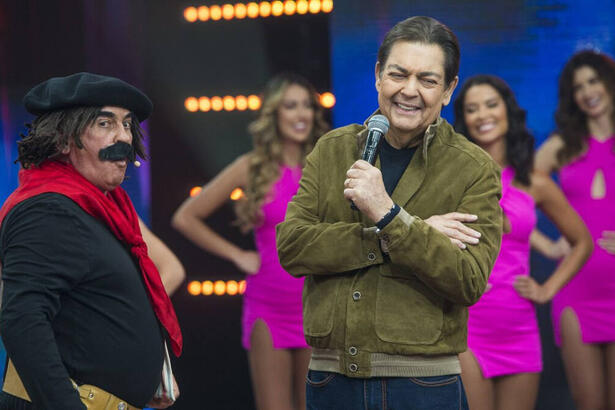 Guri de Uruguaiana com camisa preta, lenço vermelho e um bigode falso, ao lado de Faustão enquanto o apresentador sorrir durante gravação do Feira da Risada, da Band