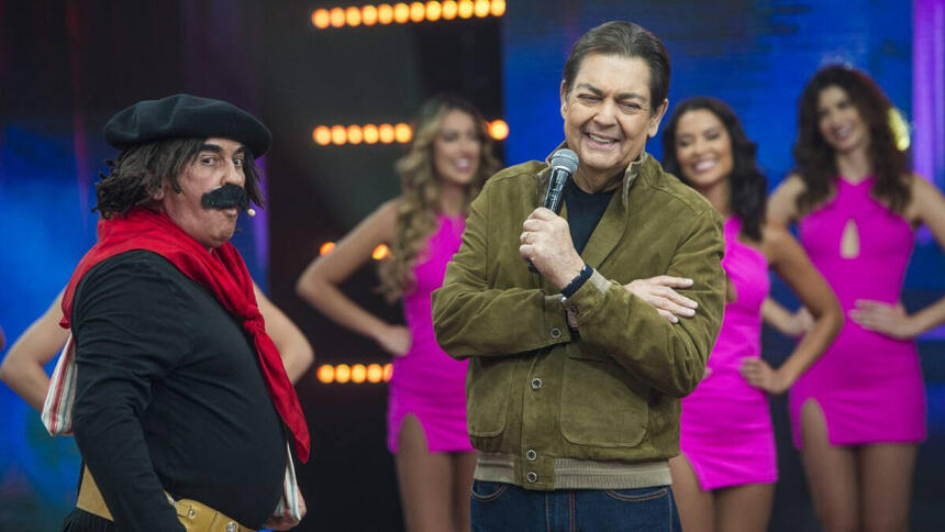 Guri de Uruguaiana com camisa preta, lenço vermelho e um bigode falso, ao lado de Faustão enquanto o apresentador sorrir durante gravação do Feira da Risada, da Band