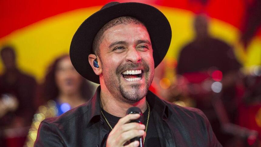 Diogo Nogueira sorrindo, de chapéu e camisa preta, segurando um microfone com a mão direita durante performance no Na Pista do Sucesso, do Faustão na Band