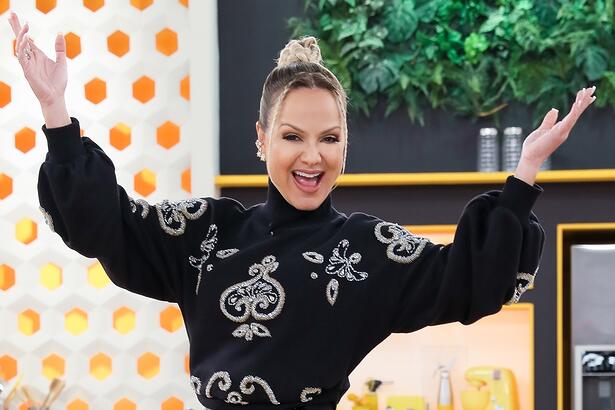 Eliana de top preto, cabelo preso, sorrindo com os braços abertos para cima durante gravação do seu programa no SBT