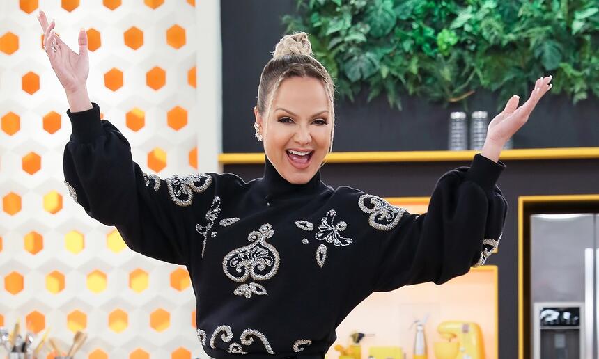 Eliana de top preto, cabelo preso, sorrindo com os braços abertos para cima durante gravação do seu programa no SBT