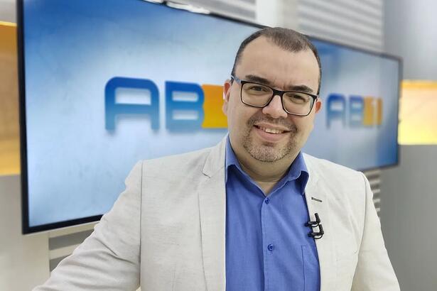 Eliaquim Oliveira de blazer cinza e camisa social azul, sorrindo, usando óculos de grau, durante apresentação do AB1 TV