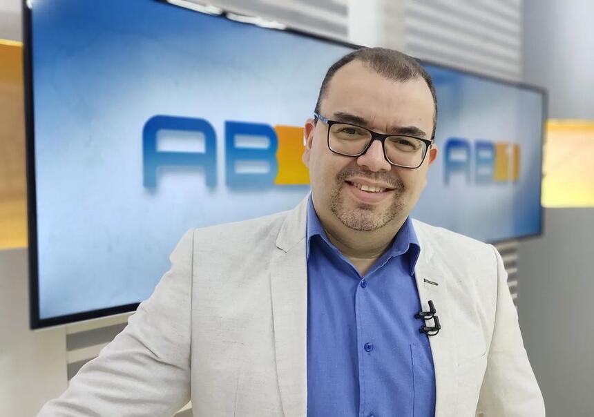 Eliaquim Oliveira de blazer cinza e camisa social azul, sorrindo, usando óculos de grau, durante apresentação do AB1 TV