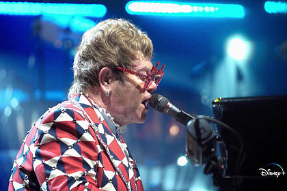 Elton John de perfil, com blazer estampado em vermelho e branco, de óculos, tocando piano durante performance em um dos seus shows bis Estados Unidos