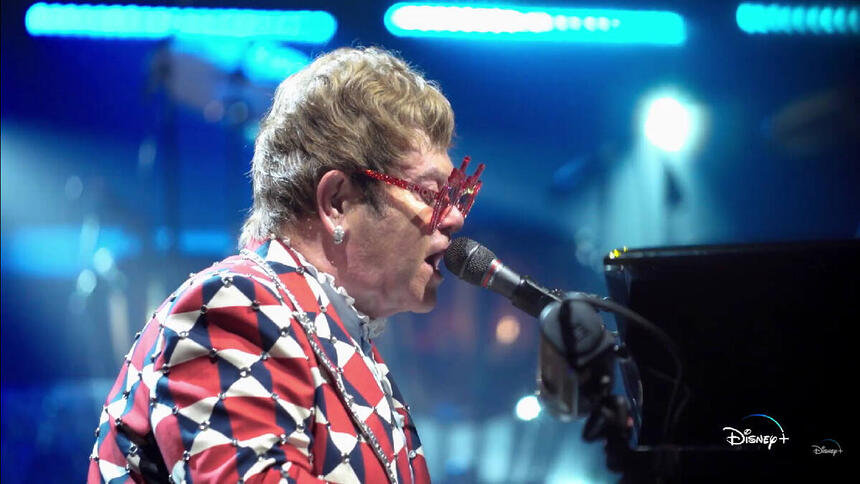 Elton John de perfil, com blazer estampado em vermelho e branco, de óculos, tocando piano durante performance em um dos seus shows bis Estados Unidos