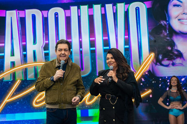 Fabiana Karla de blazer e calça pretos, sorrindo, segurando o microfone ao lado de Faustão durante gravação de programa na Band