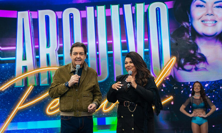 Fabiana Karla de blazer e calça pretos, sorrindo, segurando o microfone ao lado de Faustão durante gravação de programa na Band