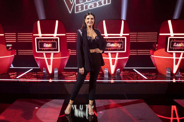 Fátima Bernardes com uma roupa preta em foto posada no estúdio do The Voice, em frente as cadeiras dos técnicos