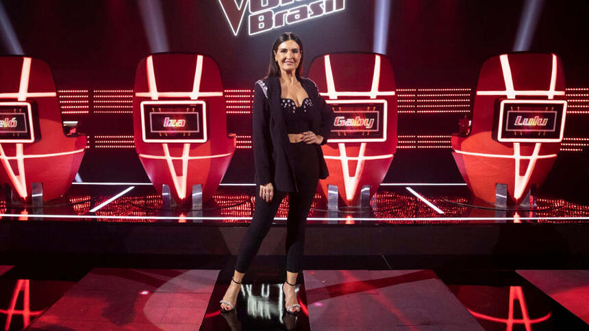 Fátima Bernardes com uma roupa preta em foto posada no estúdio do The Voice, em frente as cadeiras dos técnicos