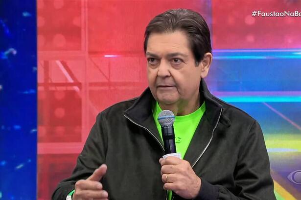 Faustão de blusa verde e casaco preto, em trecho do programa Faustão na Band, segurando o microfone com uma das mãos