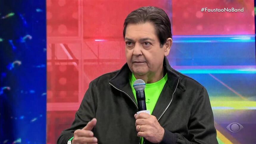 Faustão de blusa verde e casaco preto, em trecho do programa Faustão na Band, segurando o microfone com uma das mãos