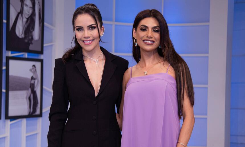 Daniela Abuquerque de vestido decotado preto, cabelo preso, sorrindo, ao lado de Flávia Noronha com vestido roxo, cabelo solto, sorrindo durante gravação do Sensacional, da RedeTV!