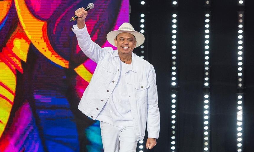 Frank Aguiar de camisa, jaqueta e calça jeans brancos, sorrindo, com o braço direito levantado durante gravação do Faustão na Band