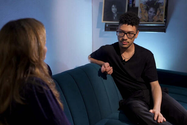 Gabriel Costa, vestido todo de preto, sentado de frente para a repórter Ana Carolina Raimundi. Eles estão em uma sala, sentados em um sofá, com quatros de Gal Costa ao fundo