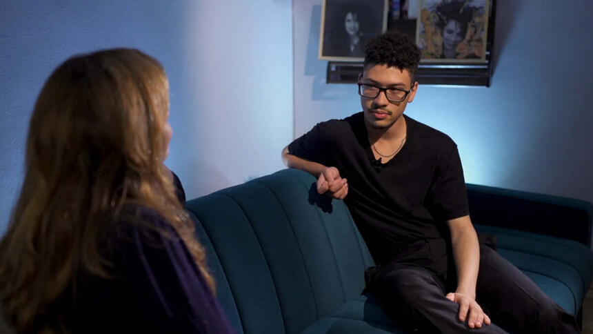 Gabriel Costa, vestido todo de preto, sentado de frente para a repórter Ana Carolina Raimundi. Eles estão em uma sala, sentados em um sofá, com quatros de Gal Costa ao fundo