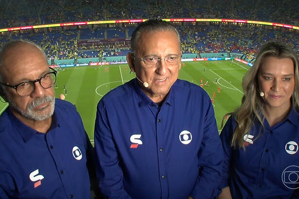 Galvão Bueno, Júnior e Ana Thaís Matos na cabine de transmissão, com o uniforme azul de esporte da Globo, com o estádio ao fundo