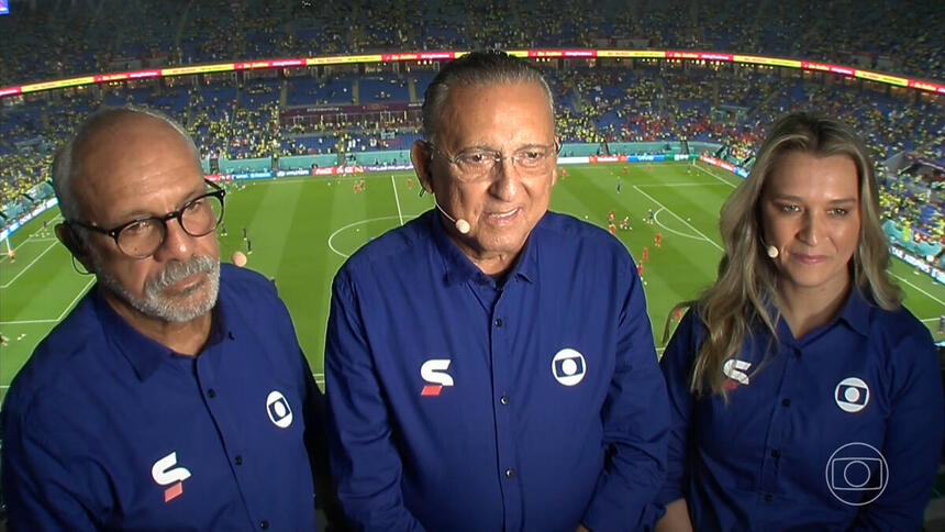 Galvão Bueno, Júnior e Ana Thaís Matos na cabine de transmissão, com o uniforme azul de esporte da Globo, com o estádio ao fundo