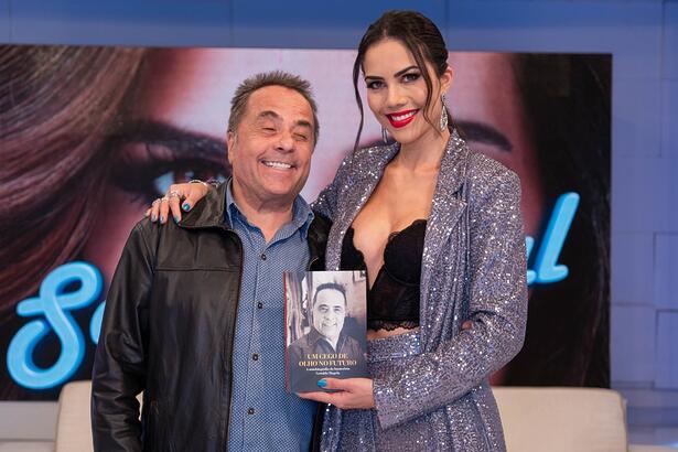 Geraldo Magela sorrindo, de jaqueta preta de couro, camisa social azul marinho ao lado de Daniela Albuquerque, que está sorrindo, segurando o livro de Geraldo, durante gravação do Sensacional, da RedeTV!