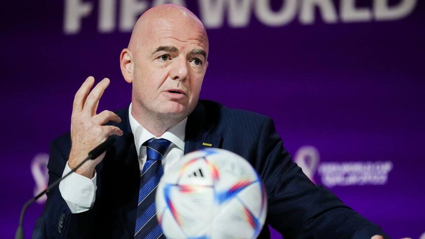 Gianni Infantino de palitó preto, gravata listrada azul e preta, com uma bola na mesa falando ao microfone durante coletiva da Copa do Mundo do Catar