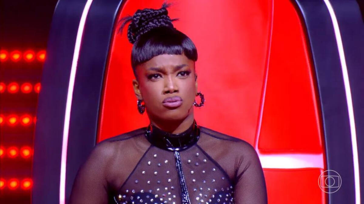IZA, com uma roupa de renda preta, sentada na cadeira de técnica, com cara de assustada, em trecho do The Voice Brasil 11