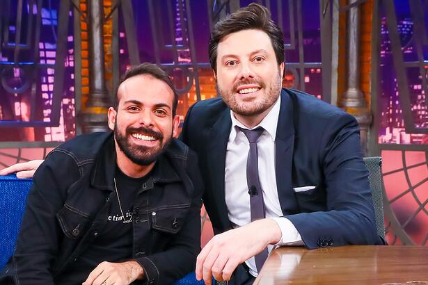 Italo Sena de camisa preta, sorrindo, sentado num sofá azul ao lado de Danilo Gentili durante gravação do The Noite, do SBT