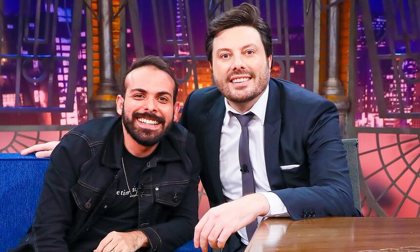 Italo Sena de camisa preta, sorrindo, sentado num sofá azul ao lado de Danilo Gentili durante gravação do The Noite, do SBT