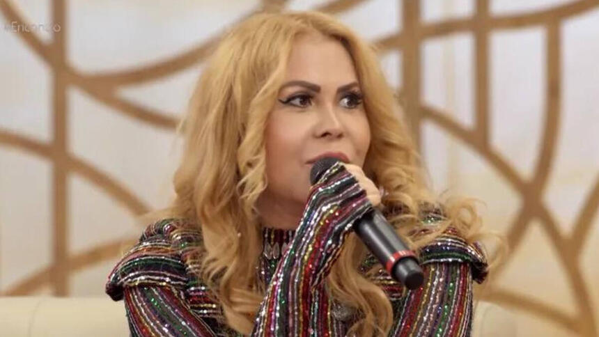 Joelma, com um microfone nas mãos, sentada no sofá do Encontro, com uma roupa brilhosa