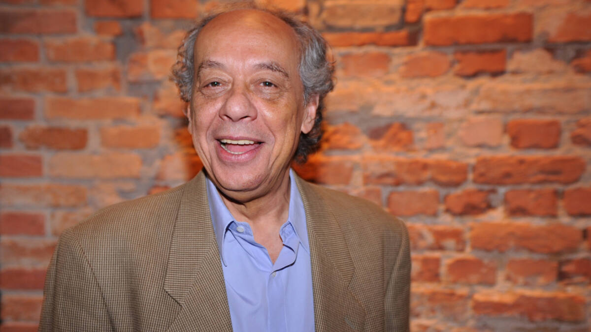 José Trajano com um blazer beje e blusa azul, em foto posada em frente a uma parede