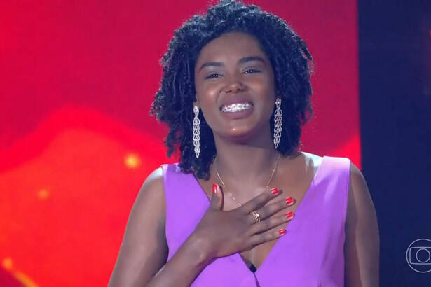 Keila Júnia com um macacão roxo, com a mão no peito, no palco do The Voice Brasil 11