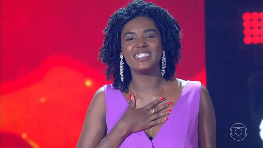 Keila Júnia com um macacão roxo, com a mão no peito, no palco do The Voice Brasil 11