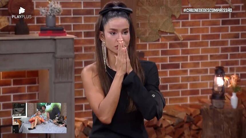 Kerline sentada com um semblante de surpresa com as mãos juntas próximo a boca.